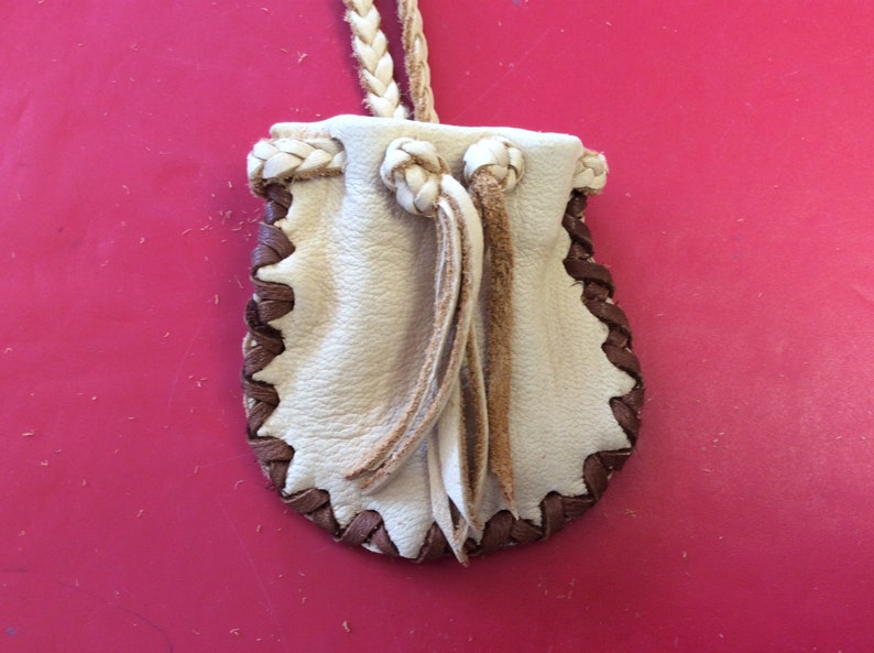 May include: A small, cream-colored leather pouch with a braided drawstring and a brown, stitched border. The pouch has a braided strap for carrying. The design is simple and rustic, suitable for small items.