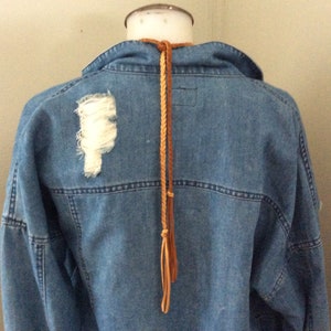 May include: A light blue denim jacket with a distressed patch on the back. A brown braided leather cord hangs from the collar. The jacket has a classic design with visible stitching.