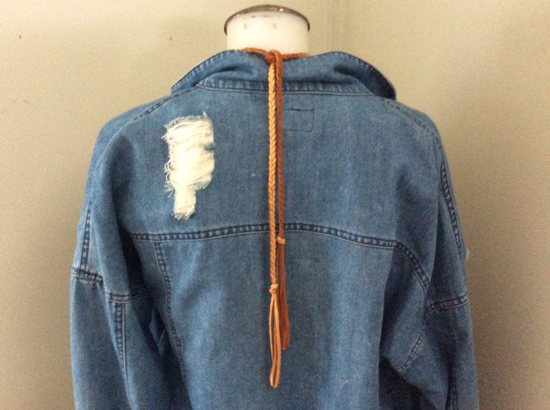 May include: A light blue denim jacket with a distressed patch on the back. A brown braided leather cord hangs from the collar. The jacket has a classic design with visible stitching.