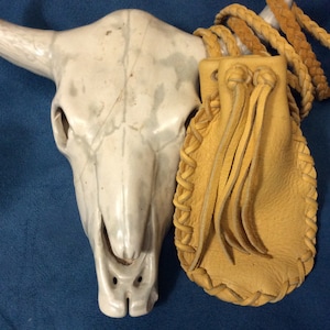 May include: A tan leather pouch with a braided leather strap and fringe. The pouch is hanging from a white ceramic bull skull.