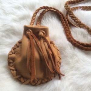 May include: A small, tan leather pouch with a braided strap and drawstring closure. The pouch has intricate braided detailing around the edges and long, fringed tassels. The bag is designed to be worn around the neck.
