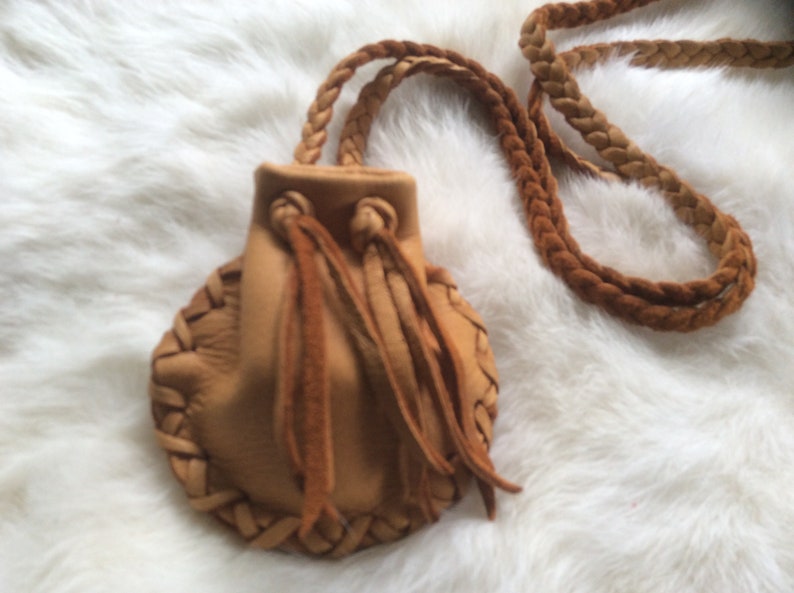 May include: A small, tan leather pouch with a braided strap and drawstring closure. The pouch has intricate braided detailing around the edges and long, fringed tassels. The bag is designed to be worn around the neck.