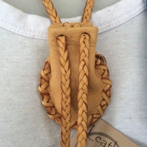 May include: A tan leather bolo tie with a braided cord and a decorative woven design. The tie is set against a white t-shirt and a denim jacket.