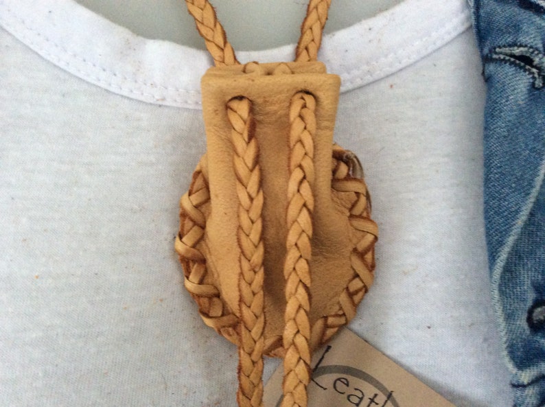 May include: A tan leather bolo tie with a braided cord and a decorative woven design. The tie is set against a white t-shirt and a denim jacket.