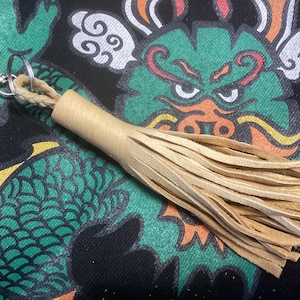 May include: A light brown leather tassel keychain with a silver-colored clasp and ring. The tassel is made of multiple strips of leather. The background features a green and black dragon design.
