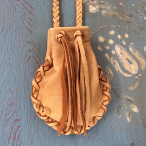 May include: A small, tan leather pouch with a braided edge and a braided strap. The pouch has a drawstring closure and decorative leather fringe. The background is a distressed blue surface.