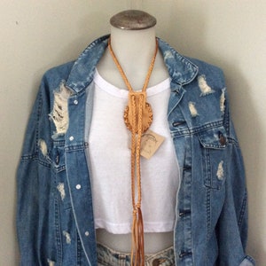 May include: A tan leather bolo tie with a circular, embossed design and braided cord. The tie is displayed on a mannequin wearing a white crop top and a distressed denim jacket. A small tag is attached to the tie.