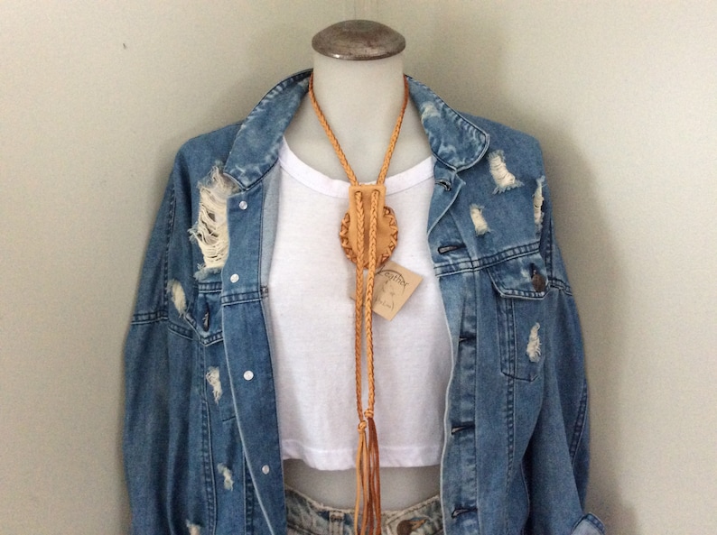 May include: A tan leather bolo tie with a circular, embossed design and braided cord. The tie is displayed on a mannequin wearing a white crop top and a distressed denim jacket. A small tag is attached to the tie.