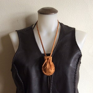 May include: A tan leather pouch necklace with a braided cord, displayed on a mannequin wearing a dark brown leather vest. The pouch has a drawstring closure and a circular design. The vest has visible stitching.
