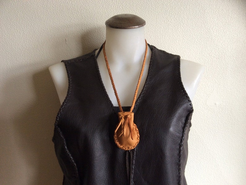 May include: A tan leather pouch necklace with a braided cord, displayed on a mannequin wearing a dark brown leather vest. The pouch has a drawstring closure and a circular design. The vest has visible stitching.