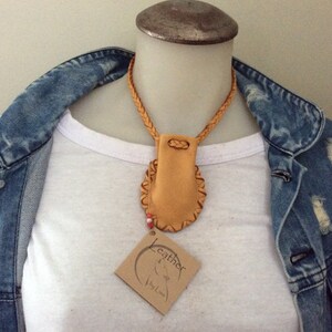 May include: A tan leather necklace with a braided cord and a small pouch pendant. The pendant has a decorative stitched border. A small tag with the word "Leather" is attached. The necklace is displayed on a white shirt and denim jacket.