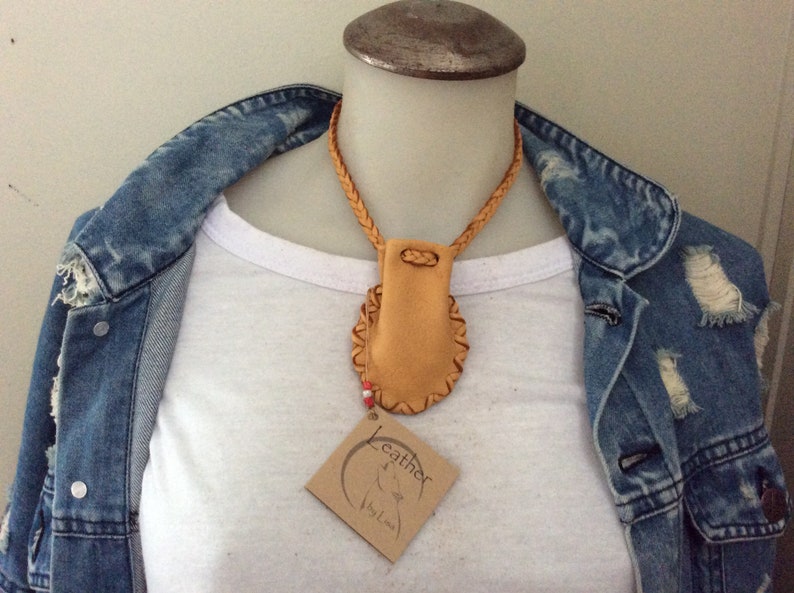 May include: A tan leather necklace with a braided cord and a small pouch pendant. The pendant has a decorative stitched border. A small tag with the word "Leather" is attached. The necklace is displayed on a white shirt and denim jacket.