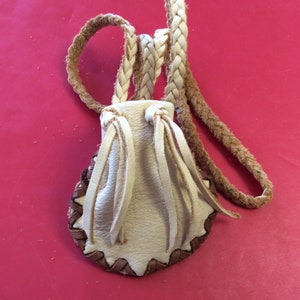 May include: A small, cream-colored leather pouch with a braided strap. The pouch has a brown, zigzag-stitched border and cream-colored fringe. The background is a solid, bright red.