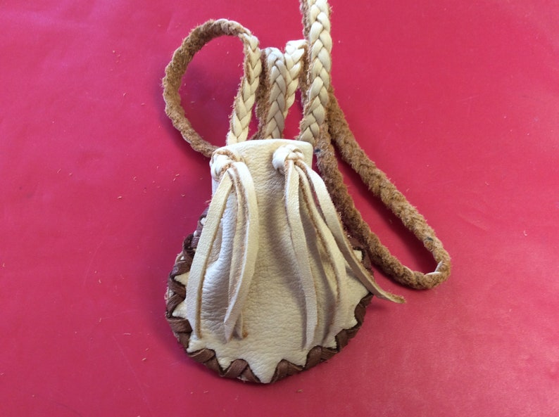 May include: A small, cream-colored leather pouch with a braided strap. The pouch has a brown, zigzag-stitched border and cream-colored fringe. The background is a solid, bright red.