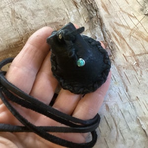 Op de afbeelding: Een klein zwart leren tasje met een turquoise steentje hangt aan een zwart leren koord.