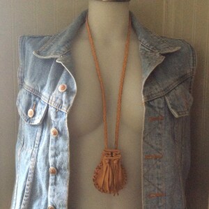 May include: A tan leather pouch necklace with a braided cord and tassel fringe. The pouch is displayed on a denim vest with copper-coloured buttons. The vest is on a mannequin.