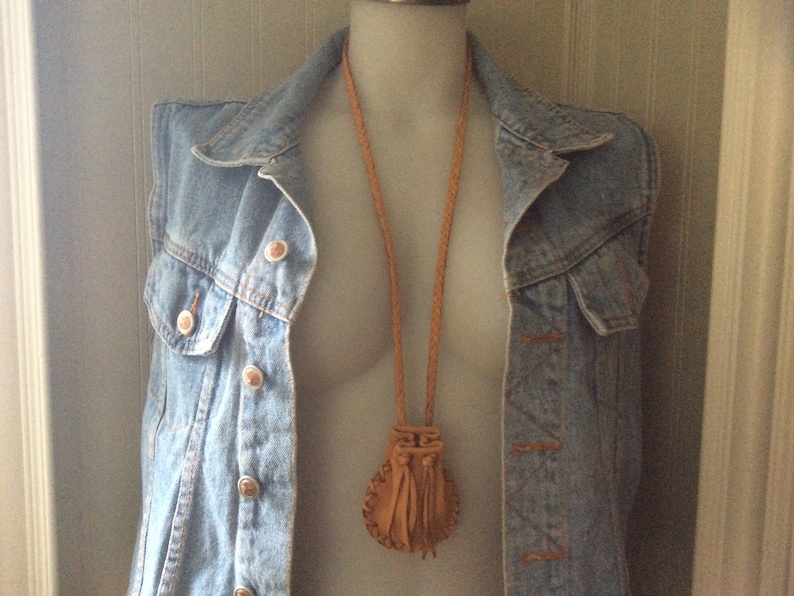 May include: A tan leather pouch necklace with a braided cord and tassel fringe. The pouch is displayed on a denim vest with copper-coloured buttons. The vest is on a mannequin.