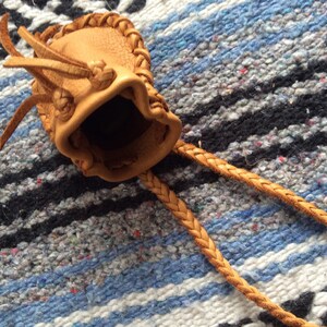 May include: A small, tan leather pouch with a braided drawstring. The pouch has a textured surface and a decorative braided trim around the opening. The drawstring is also braided and extends from the pouch.