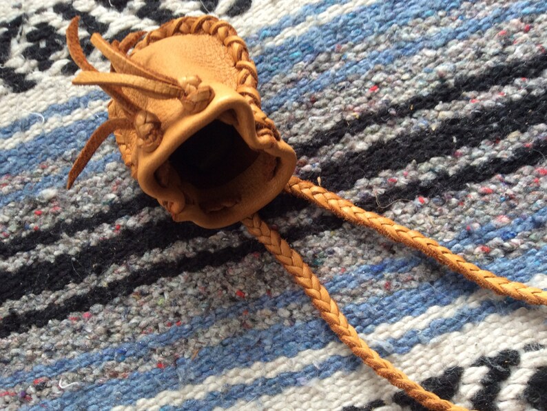 May include: A small, tan leather pouch with a braided drawstring. The pouch has a textured surface and a decorative braided trim around the opening. The drawstring is also braided and extends from the pouch.