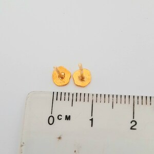 May include: A pair of gold-colored stud earrings, shown next to a ruler measuring 1 centimeter.