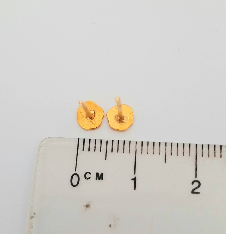 May include: A pair of gold-colored stud earrings, shown next to a ruler measuring 1 centimeter.