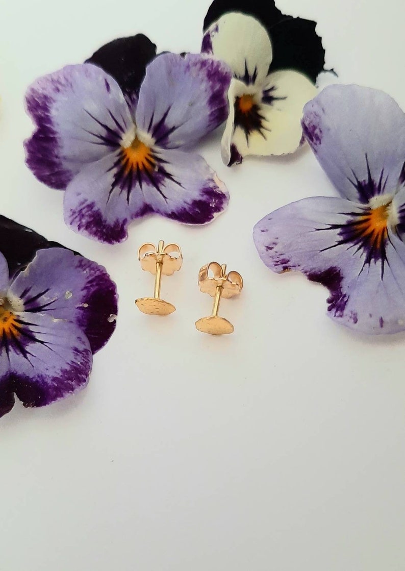 May include: A pair of gold stud earrings with a round, textured design. The earrings are displayed on a white surface with purple flowers.