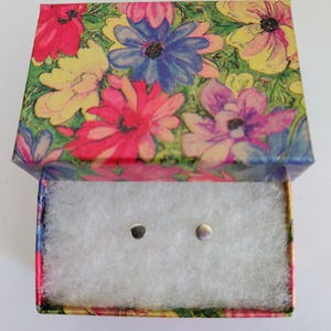 Può includere: Una piccola scatola di gioielli aperta con un motivo floreale sul coperchio. La scatola è riempita di cotone bianco e contiene due piccoli orecchini a bottone color argento. Il motivo floreale presenta fiori rosa, gialli, blu e viola.