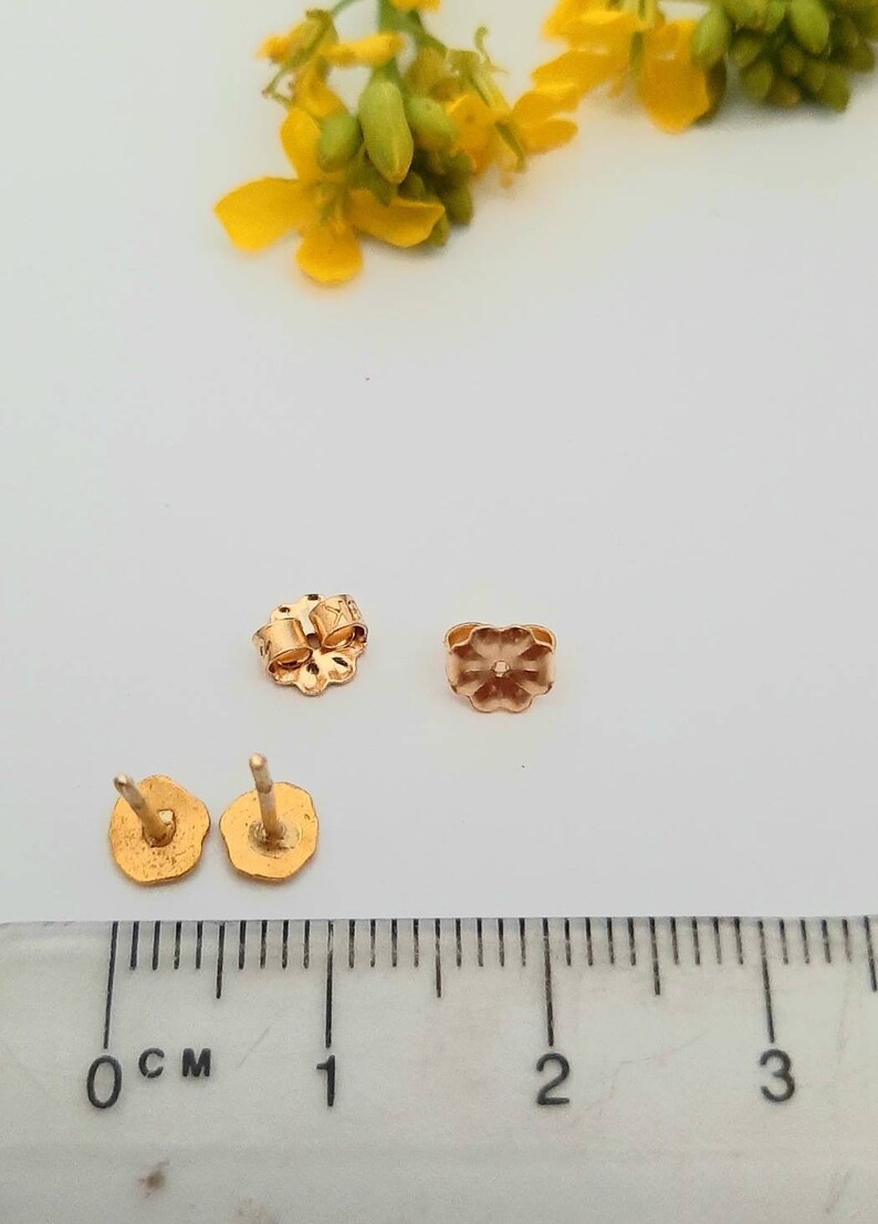 May include: Four gold-colored earring backs, two are butterfly style and two are post style, on a white surface with a ruler measuring 3 centimeters.