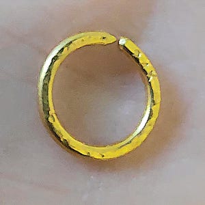 May include: A circular gold ring featuring a textured, hammered surface finish. The band is not a continuous loop, as it has a small, distinct gap at the top, creating an open-ended design. The metal has a bright, reflective yellow gold tone.