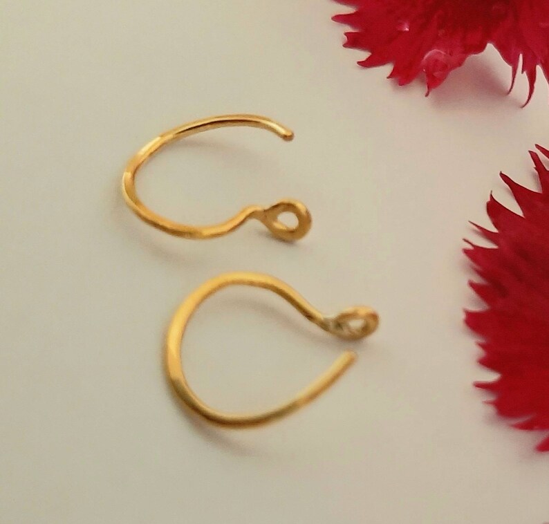 Pu&ograve; includere: Due ganci per orecchini dorati, ognuno con un piccolo anello all'estremit&agrave;. I ganci sono su una superficie bianca.