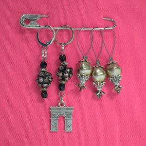 May include: A set of five silver stitch markers with black beads and a silver charm shaped like the Arc de Triomphe. The stitch markers are attached to a safety pin.