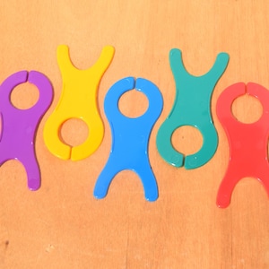 May include: Five plastic yarn bobbins in purple, yellow, blue, green, and red. Each bobbin features a central circular opening and two U-shaped cutouts at the top and bottom, designed for winding and organizing yarn or thread for various projects.