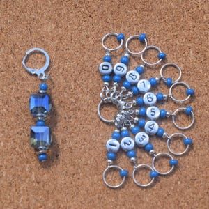 Puede incluir: Un conjunto de marcadores de punto azules y plateados para tejer. Un marcador presenta un dije de cristal azul, mientras que los otros tienen cuentas blancas numeradas y pequeños anillos plateados. Se muestran sobre una superficie marrón.