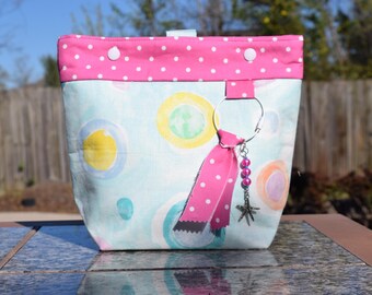 May include: A fabric tote bag with a pink polka dot top and a light blue body with colorful circles. A silver ring with a pink ribbon and starfish charm hangs from the bag.