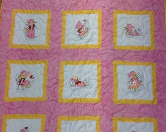 Puede incluir: Una colcha rosa y amarilla con nueve cuadrados que presentan imágenes bordadas de niñas en varias poses. Cada cuadrado tiene un fondo blanco y un borde amarillo.