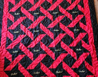 Op de afbeelding: Een rood en zwart quilt met een herhalend sterpatroon. De sterren zijn gemaakt van zwarte stof met de woorden "Crown Royal" geborduurd in goud. De quilt is gequilt met een swirl-patroon.