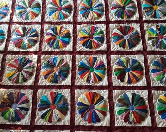 May include: A quilt featuring a grid of white square blocks, each containing a multi-colored fabric flower applique with numerous petals. The blocks are separated by a deep maroon fabric lattice, creating a structured, repeating floral pattern.