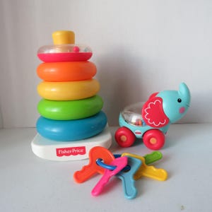 May include: A colorful Fisher-Price toy set featuring a stacking ring tower, an elephant pull toy, and a set of keys. The stacking rings are in shades of red, orange, yellow, green, and blue. The elephant is light blue with red accents.