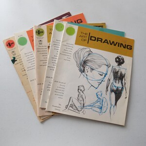 May include: A collection of five vintage instructional art books titled The Art of Drawing, featuring cream-colored covers with black ink sketches of human figures, blue construction lines, and yellow banners with black text, arranged in a fan shape.