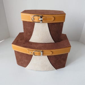 May include: Two stacked storage boxes made of brown suede with a textured finish. Each box features a central cream-colored suede panel, a mustard yellow leather-like strap with white stitching, and a silver-toned metal buckle closure.