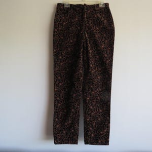 May include: A pair of brown floral print pants hanging on a white wall. The pants have a dark brown background with a repeating pattern of small, light brown and tan floral designs. The pants are straight-legged and have a button and zipper closure.