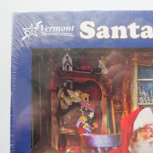 May include: A packaged jigsaw puzzle with a Christmas theme. The box features the Vermont Christmas Company logo and the word "Santa" in large white letters. The puzzle image shows Santa Claus in a room with a fireplace and toys.