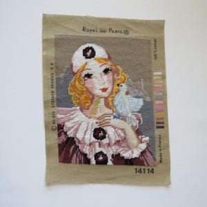 May include: Needlepoint canvas featuring a vintage illustration of a woman with blonde hair and a white hat, holding a blue bird. The artwork is stitched with various colors and has the text "Royal Paris" and "Made in France".