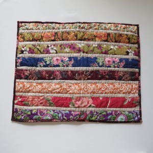 May include: A rectangular quilt with horizontal stripes of various floral patterns in shades of orange, pink, blue, and purple. The quilt has a dark brown border and is likely handmade, showcasing a patchwork design.