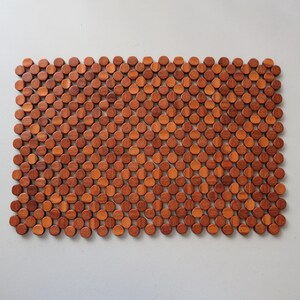 May include: A rectangular mat constructed from numerous small, circular wooden discs arranged in a grid pattern. The discs feature varying shades of warm brown, ranging from light tan to deep reddish-brown, creating a textured, honeycombed surface.