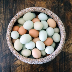 May include: A woven basket filled with a variety of fresh eggs. The eggs are in shades of white, light blue, and peach. The basket has a decorative woven pattern around the rim, and sits on a dark wooden surface.