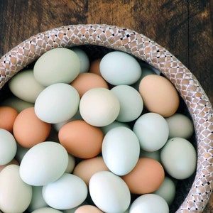 May include: A woven basket filled with a variety of fresh eggs. The eggs are in shades of light blue, tan, and cream. The basket has a woven pattern and sits on a dark wooden surface.