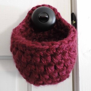 May include: A burgundy crocheted hanging basket with a black button closure. The basket is hanging on a white door.
