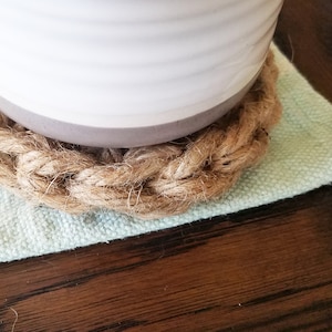 May include: A close-up of a woven jute coaster with a white ceramic pot on top. The coaster is on a light green fabric surface.