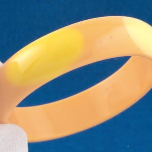 Op de afbeelding: Een gladde, ronde armband met een effen perzikkleurige basis en een opvallend, langwerpig felgeel ovaal accent op de buitenkant. De armband heeft een glanzende, gepolijste afwerking en een eenvoudig, doorlopend cirkelvormig ontwerp.